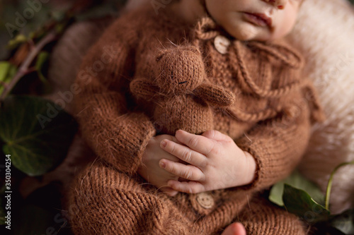 A newborn charming boy, 5 days old, sleeps in a cozy bear outfit and in a light brown outfit. He sleeps in a basket and holds a tiny teddy bear.