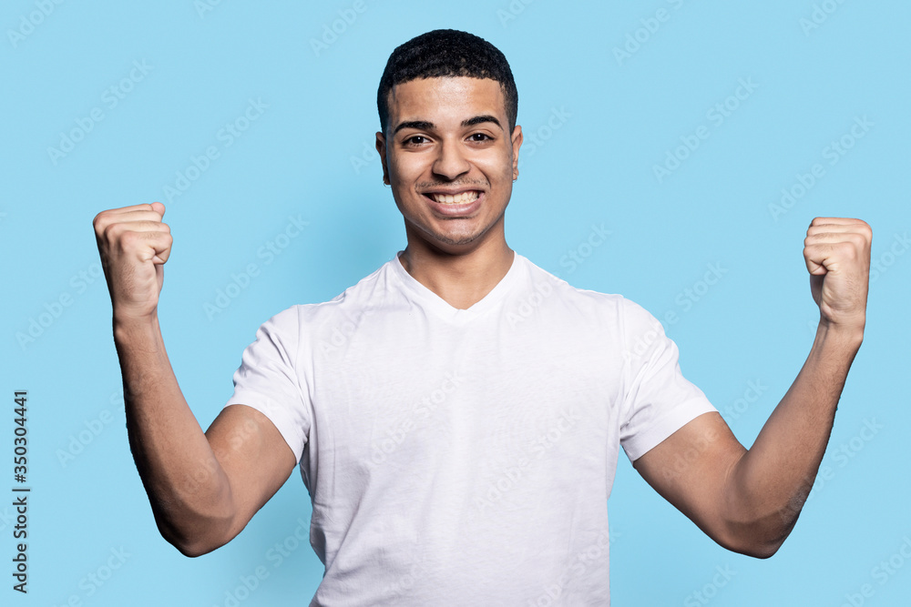Fototapeta premium Overjoyed black man raises clenched fists, feels energized while gesturing happily against blue wall