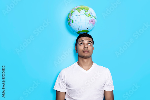 Young black male standing against blue wall while holding earth globe with his head and looking up