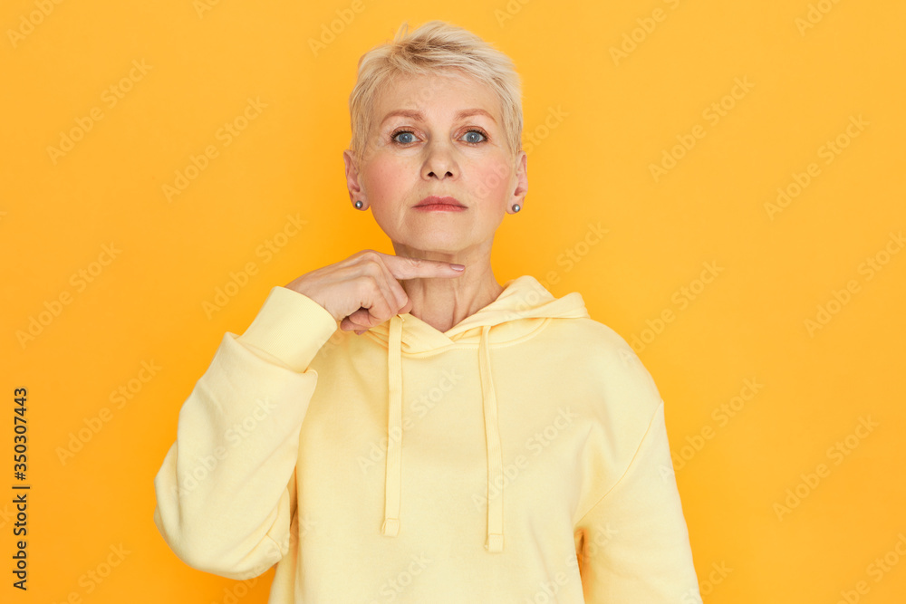 Serious Caucasian mature woman in stylish hoodie, making threatening sign, gesturing with index finger at her neck as if cutting throat. Negative human facial expressions and body language
