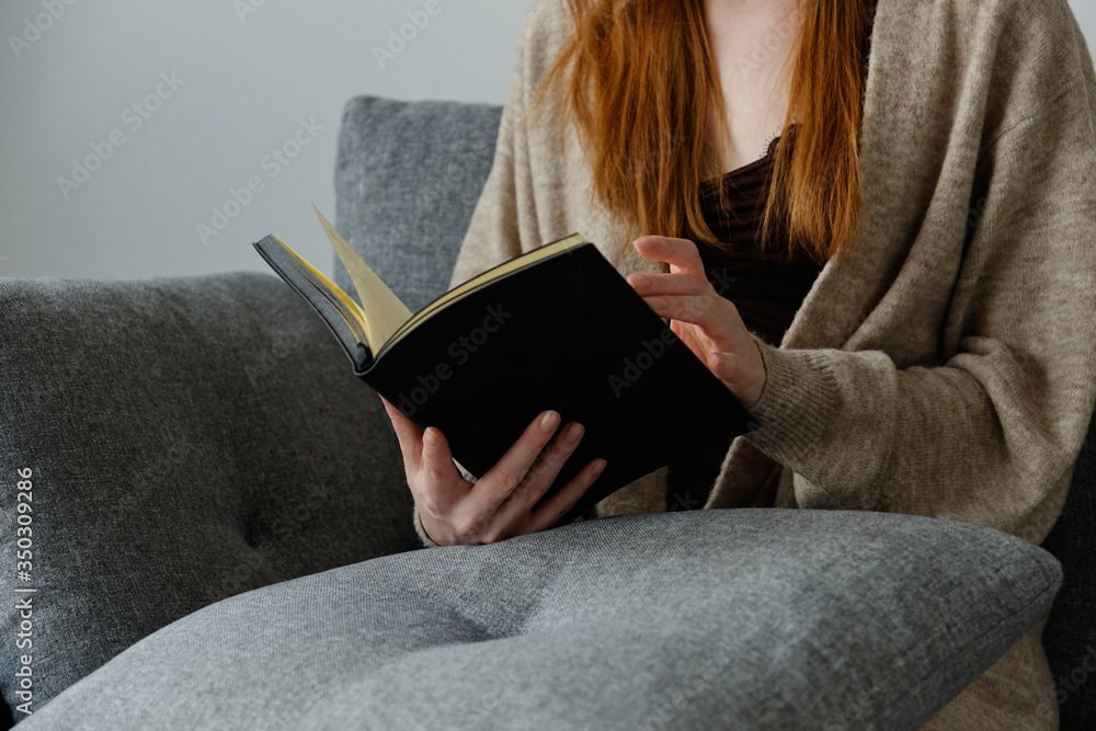 Obraz premium A black book in the hands of a red-haired girl sitting on a sofa.