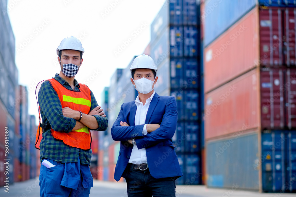 Technician or engineer workers stand in front of shipping container with one hold infrared thermometer and manager also wear face mask in the procedure to prevent virus infection in workplace area. Technician or engineer workers stand in front of shipping container with one hold infrared thermometer and manager also wear face mask in the procedure to prevent virus infection in workplace area.
