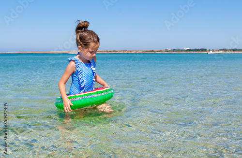 Happy cute tollder girl 4 or 5 years old in a bright blue swimsuit with swim tubes ring goes into the sea for swimming on a sunny day. Family vacation with children concept photo