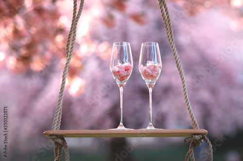 Two glasses stand on a wooden swing in a flowering spring garden.