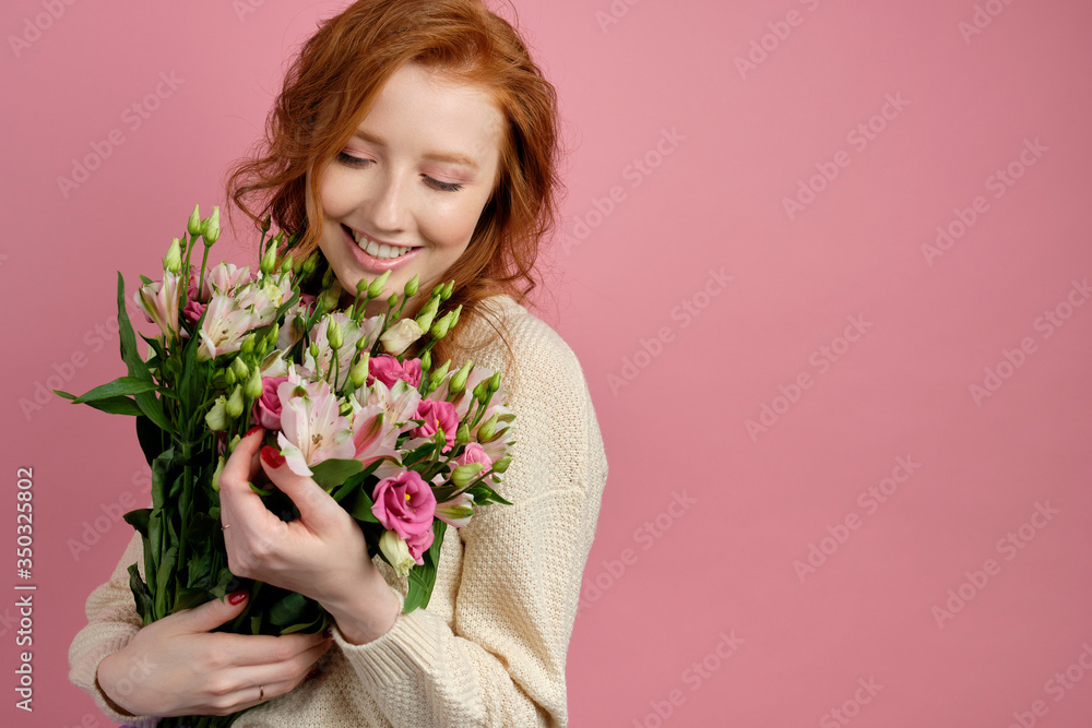 Cute red-haired girl stands on a pink background, with a smile presses a bouquet to herself, looking at him Cute red-haired girl stands on a pink background, with a smile presses a bouquet to herself, looking at him