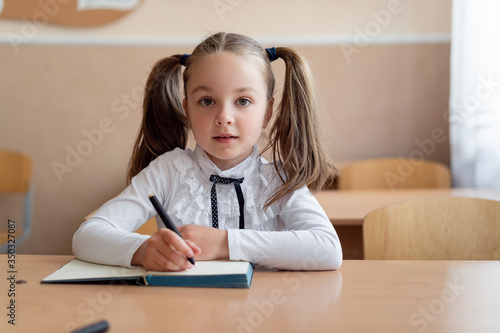 little girl schoolgirl is sitting at a desk. Hairstyle two braids on the head. Hold a pen and look at the teacher. Write in a notebook.