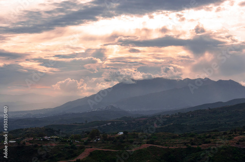 Bright light breaks through the clouds during sunset over the mountain peaks, a country road, green hills and white private houses in the foreground.