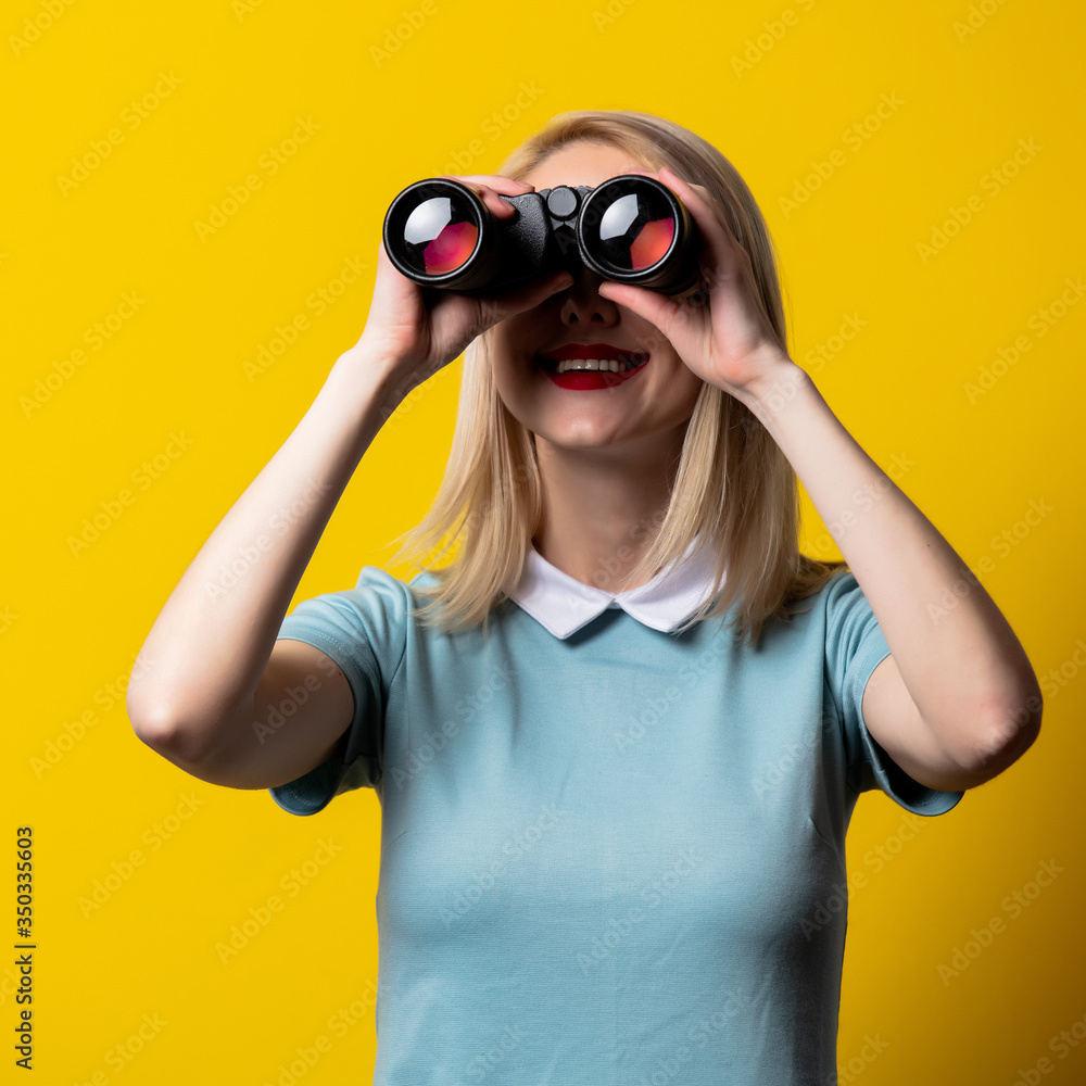 Blonde girl in blue dress watching in binoculars on yellow background Blonde girl in blue dress watching in binoculars on yellow background