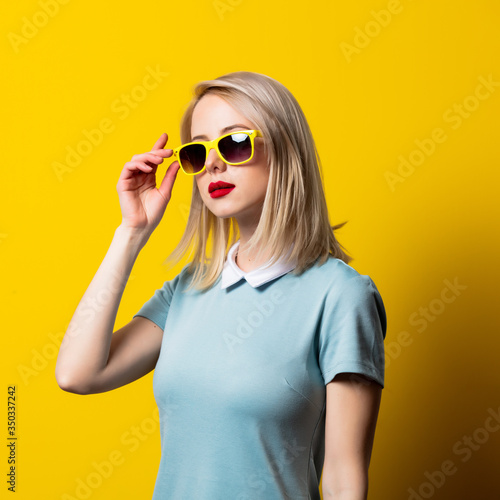 Blonde girl in sunglasses and blue dress on yellow background