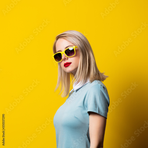 Blonde girl in sunglasses and blue dress on yellow background