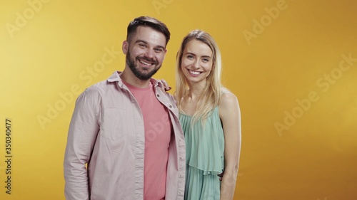 Cute caucasian young couple standing and posing on orange background. Their funny bearded friends appearing behind backs smiling happy together. Copy space. Friendship.