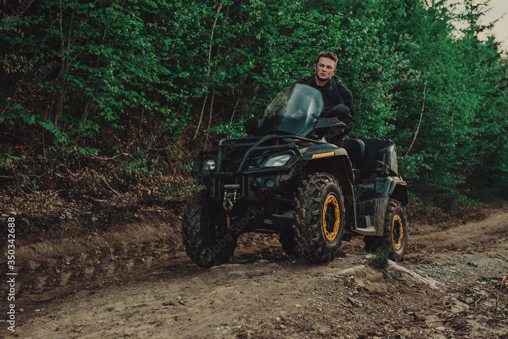 Fototapeta premium A young man in a white helmet rides through the woods on a Quad bike. Extreme hobby. A trip to ATV on the road from logs. Quad Biking through the forest.