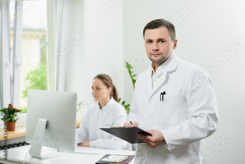A serious surgeon with bristles in a white coat holds a black clipboard with documents that stays in a hospital. A female doctor sitting in front of the desktop computer in the corner of the room. 