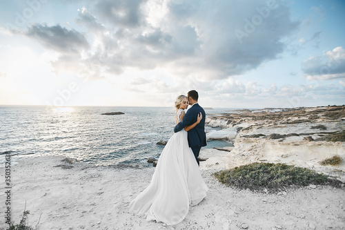Newlywed couple hugging together on the white rocks at the seacoast at Cyprus. The bride in a wedding dress the groom in a suit posing with beautiful landscape on the background
