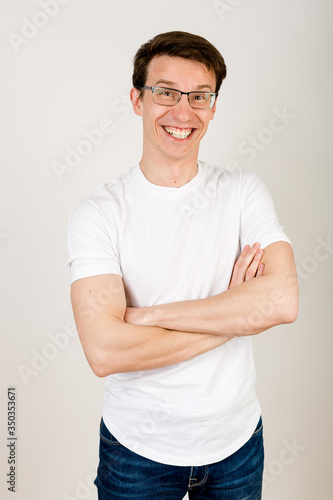 A young hipster in casual clothes stands on a white background with his hands folded on his chest and laughs heavily, looking into the camera.