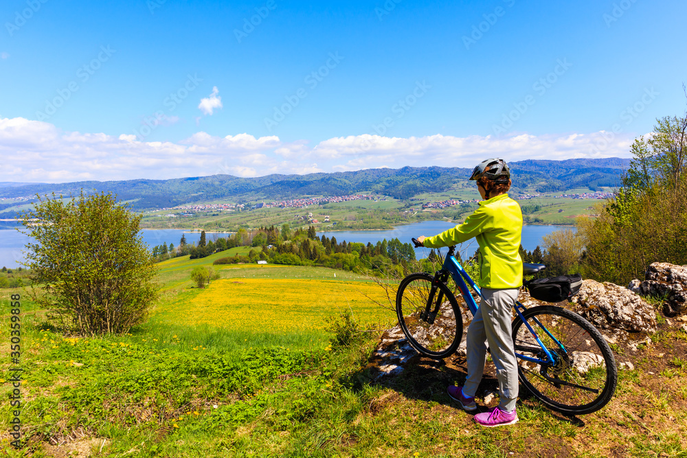 Woman cyclist standing on a rock and looking at Czorsztynskie lake near Falsztyn village on sunny spring day, Pieniny Mountains, Poland