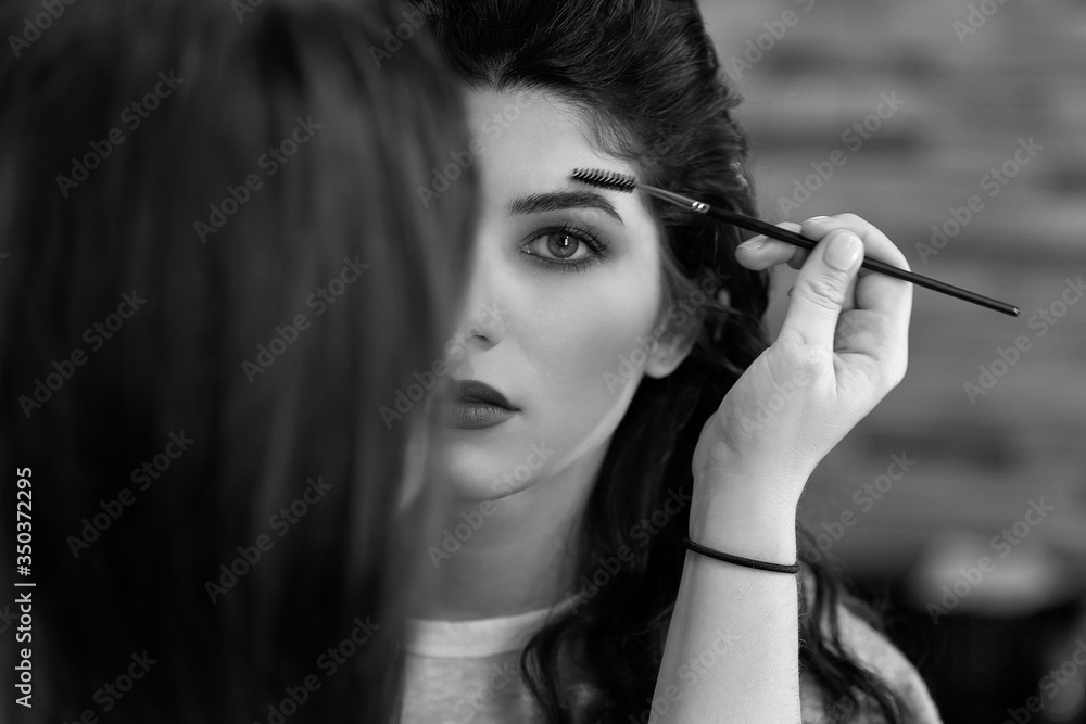 Obraz premium Black and white portrait of a young girl with dark long hair, whom the stylist does makeup, combing her eyebrows with a brush. Makeup model before photo shoot in the studio. Makeup before fashion show