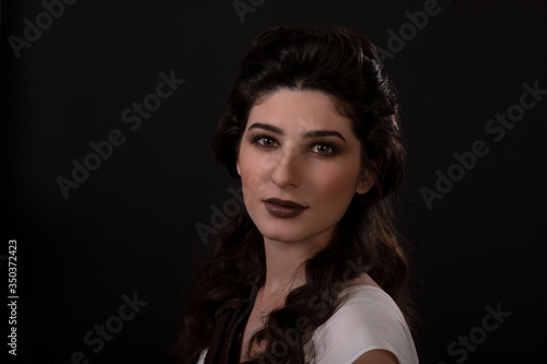 Close-up portrait of a archer woman in white dress on black background. Greek goddess Artemis. Studio photo shoot. Professional makeup and hairstyle for a brunette with long dark hair. Young woman