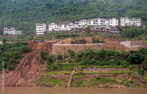 Fengdu, Chongqing, China - May 8, 2010: Yangtze River. White housing row near green forest on top of shoreline where colapsed road is being repaired with trucks and cranes present. Brown water. photo