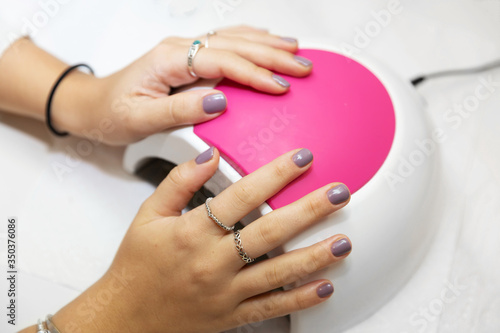 Ready manicure in purple. Hands of a young girl with a gentle purple manicure on a machine for ultraviolet drying gel varnish in the salon. Nail care after quarantine.