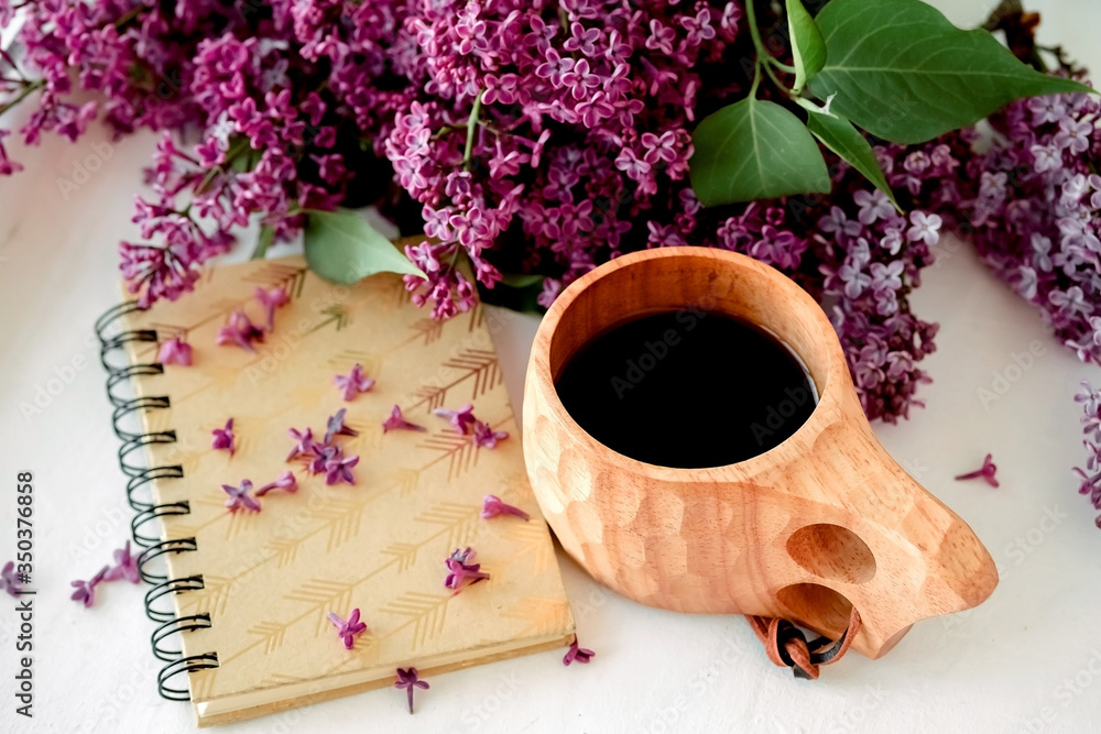 Wooden finnish cup with coffee on the table. Nearby lies a closed notebook for notes and a bouquet of lilacs. Good morning and good mood, planning a day at breakfast, concept. Wooden finnish cup with coffee on the table. Nearby lies a closed notebook for notes and a bouquet of lilacs. Good morning and good mood, planning a day at breakfast, concept.