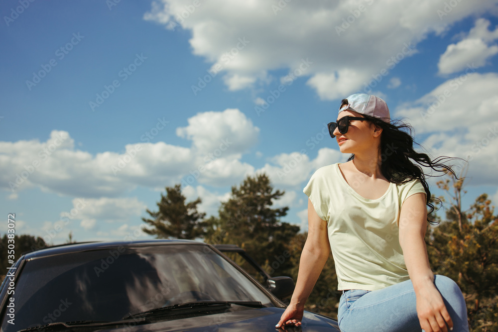 Young woman traveler sitting on the car hood while traveling. Road trip, vacation and adventures