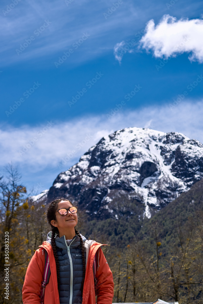 Beautiful and colorful portrait in a cold winter of a smiling beautiful young woman wearing orange jacket and sunglasses. Surrounded by a dry forest with snow capped mountains behind and a blue sky