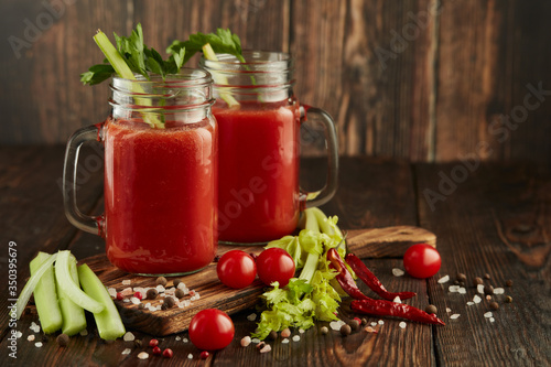 Two glasses with fresh tomato juice, celery, parsley and ripe tomatoes on dark brown wooden background.