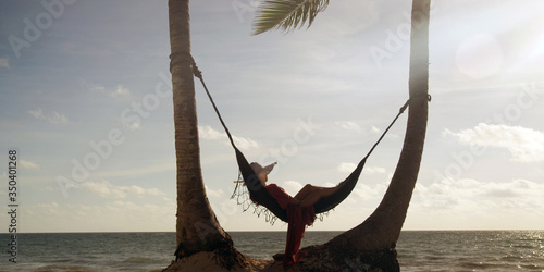 Woman rests in loose red summer dress and hat hanging in hammock surrounded by palm trees at sunset