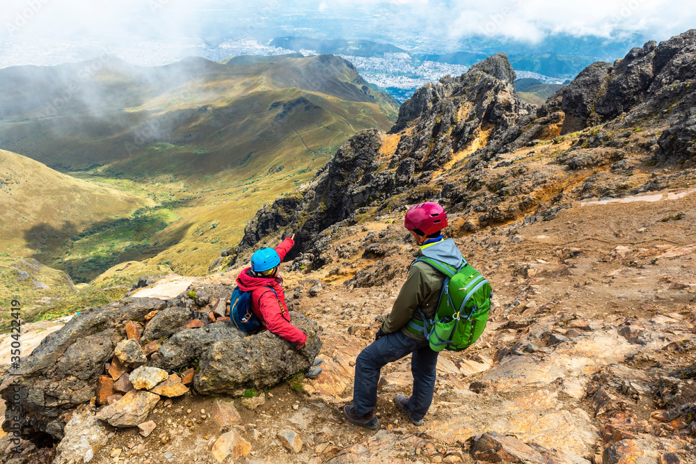 Two young backpackers with protection helmet along the Rucu Pichincha Volcano hike and with the aerial view over Quito, Andes mountains, Ecuador.