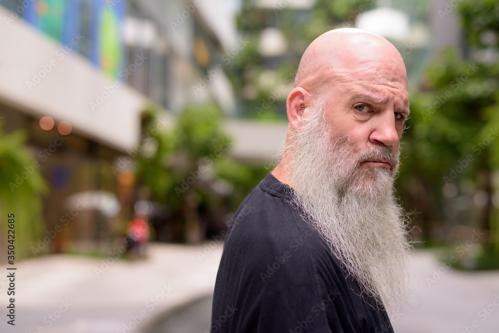 Face of mature bald bearded man looking serious at camera with nature in the city Face of mature bald bearded man looking serious at camera with nature in the city