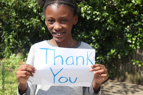 Kid smiling while holding Thank You placard sign outside with green bushes in background