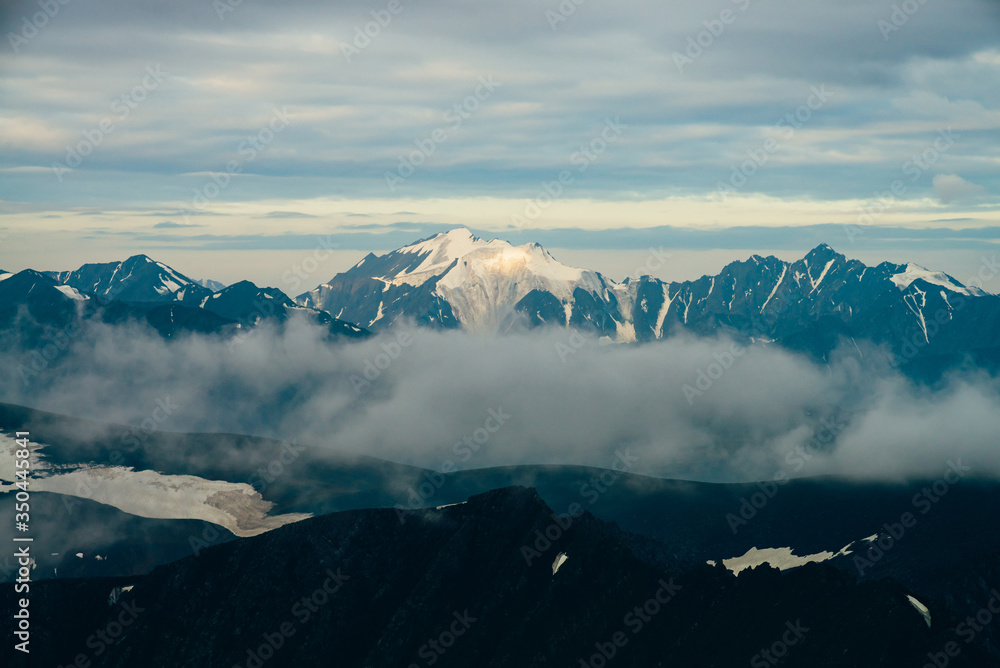 Atmospheric alpine landscape with big snowy mountains among low clouds in golden hour. Wonderful highland scenery with massive glacier on giant mountain range in sunrise. Shiny snow on big rockies.