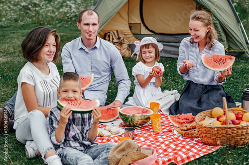 Happy family eating watermelon at picnic in meadow near the tent. Family Enjoying Camping Holiday In Countryside