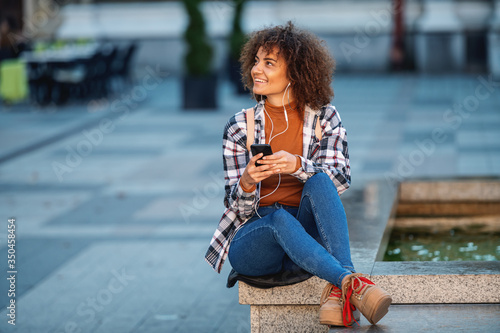 Young attractive mixed race woman sitting on fountain, using smart phone and enjoying music.