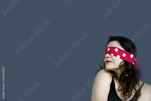 Portrait of a beautiful, fashionable middle-aged woman with long dark hair and a red blindfold covering her eyes, posing against a dark gray background.