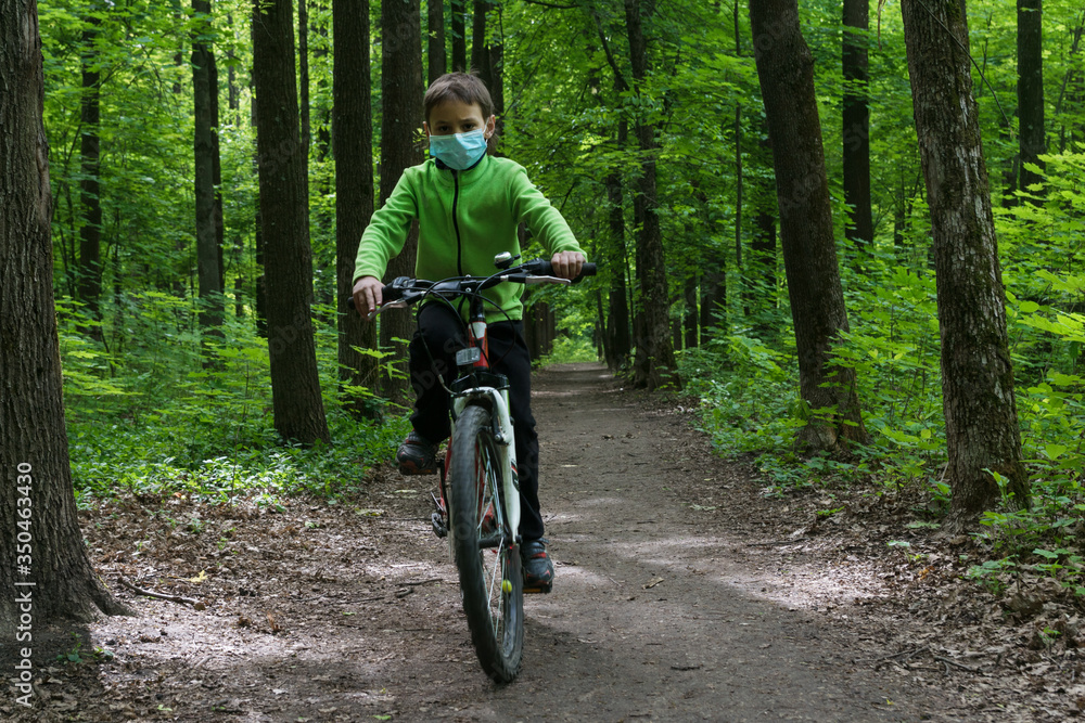 boy in protective mask rides a bike in deep green forest, safe new way of sport activities after end of quarantine lockdown, outdoor sport activities
