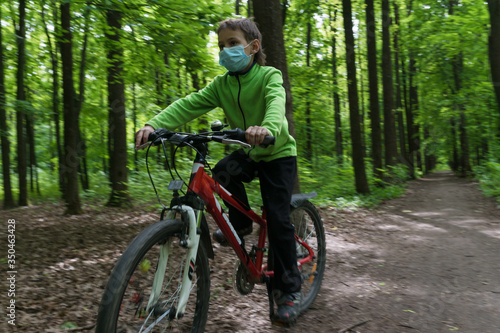 boy in protective medical mask rides a bike in deep green summer forest, safe new way of sport activities after end of quarantine lockdown, outdoor sport activities © Anna