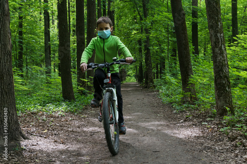 boy in protective mask rides a bike in deep green forest, safe new way of sport activities after end of quarantine lockdown, outdoor sport activities