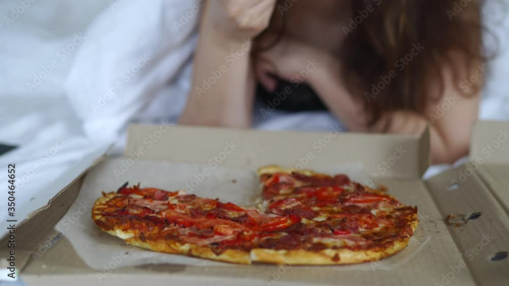 Woman eating pizza in bed, pizza served in a cardboard box, fast food delivery. Woman eating pizza in bed, pizza served in a cardboard box, fast food delivery.