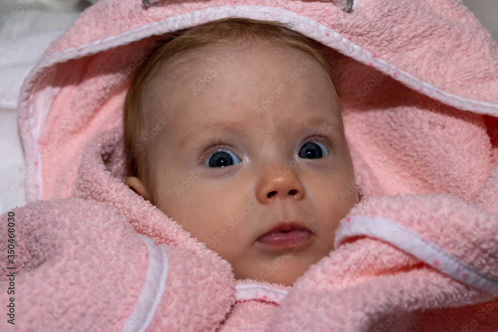 Portrait of Infant baby girl under the pink towel roll looking at camera lying on bed after bath. 3 months old baby girl. Portrait of Infant baby girl under the pink towel roll looking at camera lying on bed after bath. 3 months old baby girl.
