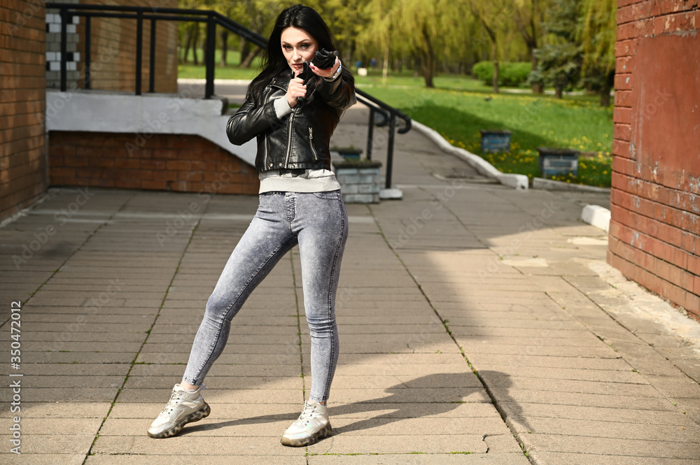 Full length portrait of a caucasian fashionable brunette girl in black jacket and jeans standing outdoors in springtime outdoors. Model posing against the backdrop of a city park with an umbrella.
