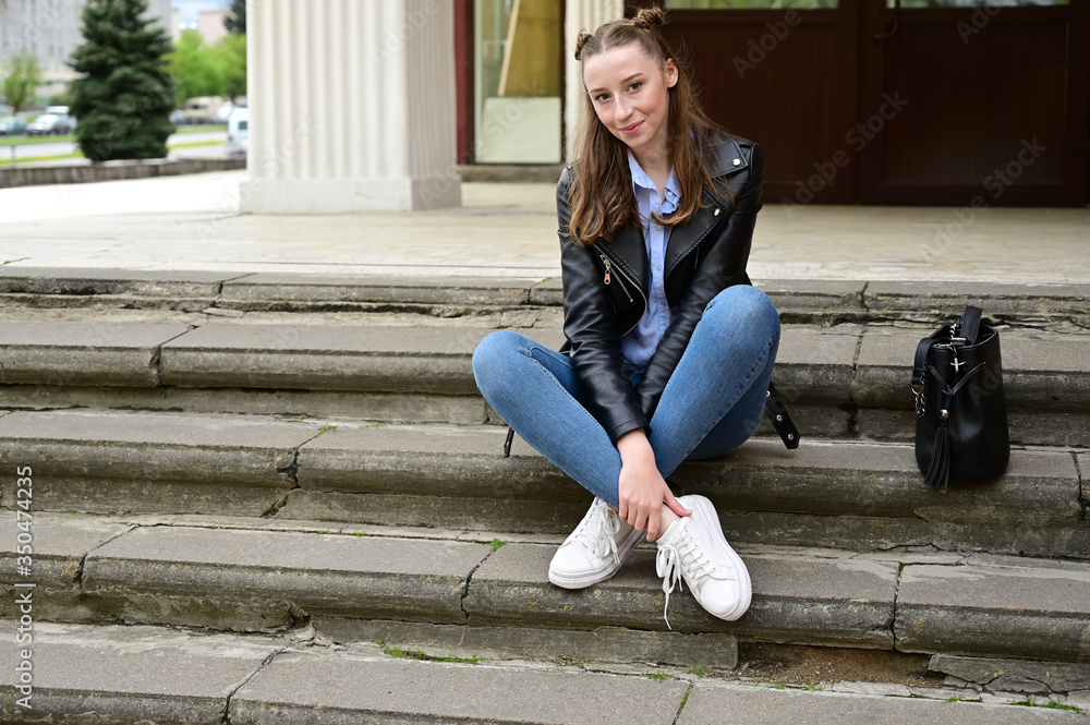 Model posing sitting in spring park outdoors in the city on the steps. Photo of a young pretty girl with a smile in a black jacket and jeans.