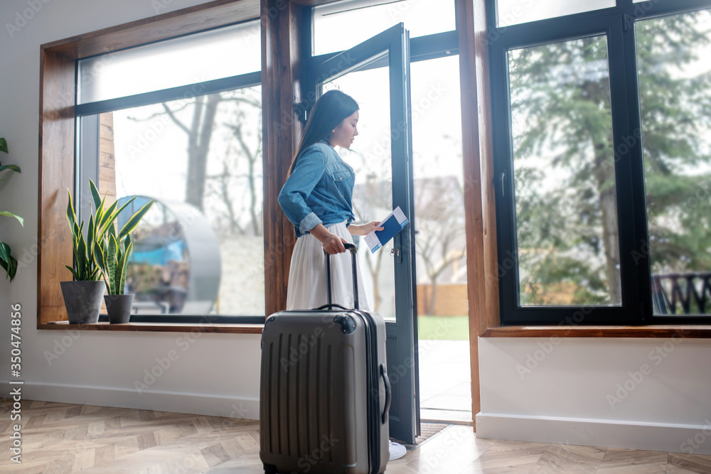 Dark-haired female opening glass door, pulling her suitcase, leaving