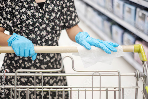 Asian woman wearing nitrile gloves and cleaning surface of shopping cart by wet wipe in supermarket or grocery, protect coronavirus inflection. Hygiene, new normal and life after covid-19 pandemic