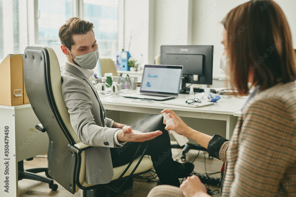 Woman spraying sanitizer onto colleagues hands while they working in half-empty office Woman spraying sanitizer onto colleagues hands while they working in half-empty office