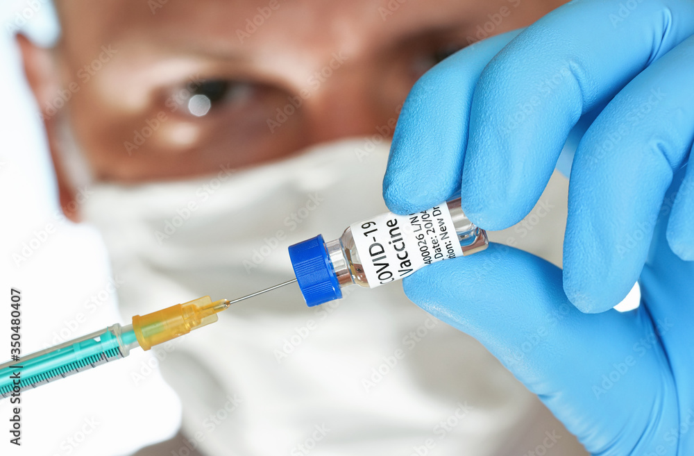 Man hand in blue nitrile glove hold small vial with coronavirus vaccine, to inject orange syringe, blurred face with white virus mask in background (sticker is own design, dummy data not real product) Man hand in blue nitrile glove hold small vial with coronavirus vaccine, to inject orange syringe, blurred face with white virus mask in background (sticker is own design, dummy data not real product)