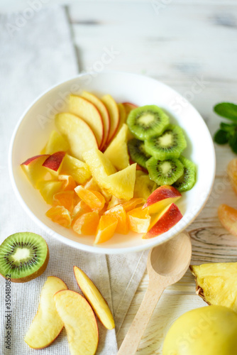 Close up view of sliced fruits in a white bowl and wooden spoon with fruits around on a bright background