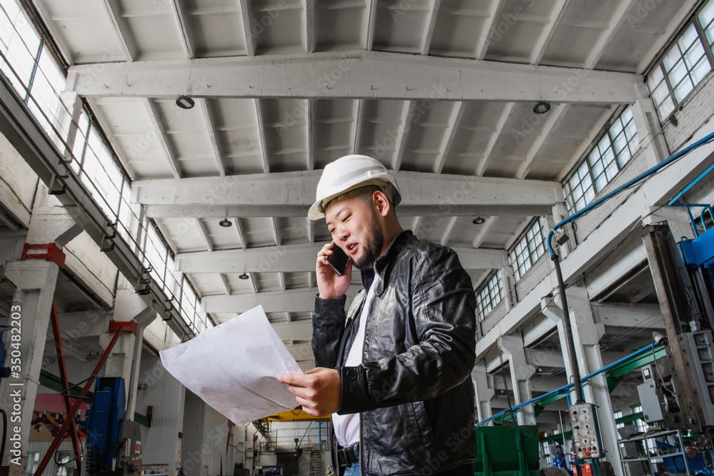 Young Asian engineer in a modern factory, holding a drawing in his hands and talking on the phone. The photo illustrates new technologies and production.