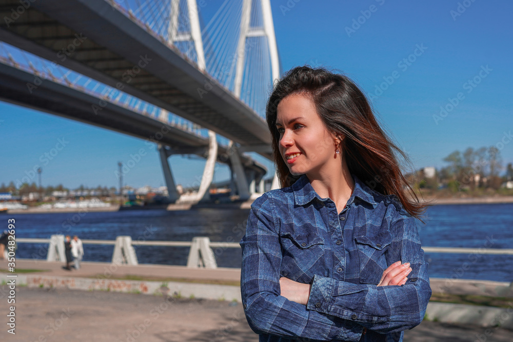 A brunette girl with long hair in a blue shirt stands against the background of a cable stayed bridge on the embankment A brunette girl with long hair in a blue shirt stands against the background of a cable stayed bridge on the embankment